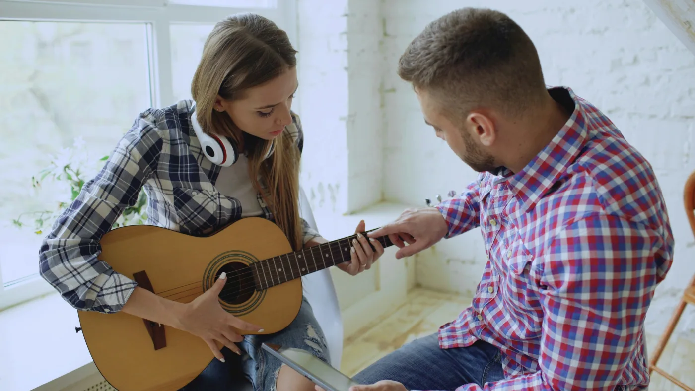 A guitar teacher pointing at the fretboard as a student plays an acoustic guitar by a bright window.