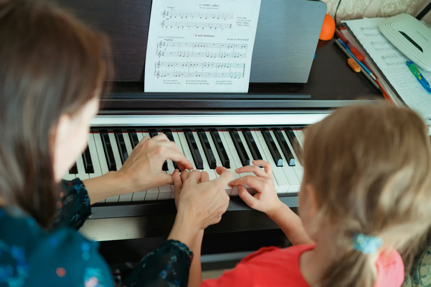A teacher's hands guiding a young student's hands on the piano keys, sheet music on the stand.
